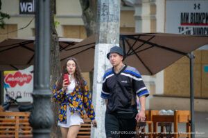 Фото мужчины с татуировками на руке и шее, идущего с девушкой по улице. Мужчина курит сигарету, девушка смотрит в телефон.