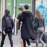 Фотография группы людей, идущих по улице. Видна татуировка на руке одной из девушек. Городской пейзаж, яркий день.