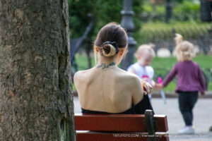 Фото тату: девушка с нежной татуировкой на задней части шеи сидит на скамейке в парке. Рисунок выполнен в черных тонах.