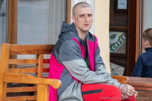 Фото тату на лице и руках мужчины, сидящего на скамейке возле кафе. Видны надписи и узоры на коже, отражающие современный уличный стиль.