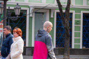 Фотография мужчины с короткой стрижкой и татуировкой в виде креста на шее. Он одет в яркую розово-серую куртку и смотрит в сторону.