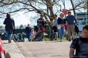 Группа людей прогуливается по улице в солнечный день. У одного из мужчин видны татуировки на руках и ногах.