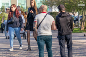 Фотография группы людей, идущих по улице. У одного из мужчин видна татуировка на руке. Современный городской пейзаж.