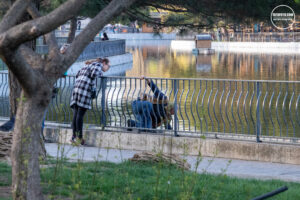 Фотография двух девушек, кормящих голубей у пруда. Одна стоит на мосту, другая наклонилась к воде. Яркий солнечный день.