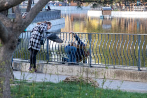 Фотография двух девушек, кормящих голубей у пруда. Одна стоит на мосту, другая наклонилась к воде. Яркий солнечный день.
