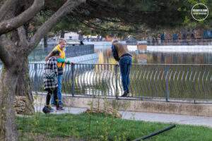 Фотография группы людей, стоящих у перил и смотрящих на воду. Видна река или озеро и отражение неба в воде.