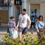 Фото мужчины с татуировками на руках и в футболке с ярким принтом. Татуировки выполнены в черном цвете, а принт на футболке сочетает желтый, черный и белый цвета.