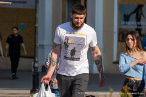 Фото мужчины с татуировками на руках и в футболке с ярким принтом. Татуировки выполнены в черном цвете, а принт на футболке сочетает желтый, черный и белый цвета.