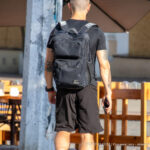 Фото тату на спине мужчины, идущего по улице с черным рюкзаком. Видна часть татуировки, выполненной в темных тонах.