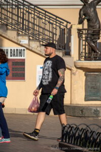 Фотография мужчины с татуировкой на руке, идущего по улице. Виден яркий и детализированный рисунок с изображением волка и других элементов. Снимок сделан на фоне скульптуры.