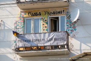 Фотография фасада дома, украшенного яркими рисунками и надписями в поддержку Украины. На балконе висят флаги и плакаты.