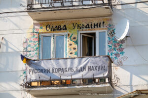 Фотография фасада дома, украшенного яркими рисунками и надписями в поддержку Украины. На балконе висят флаги и плакаты.