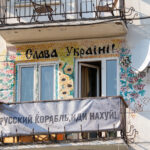 Фотография фасада дома, украшенного яркими рисунками и надписями в поддержку Украины. На балконе висят флаги и плакаты.