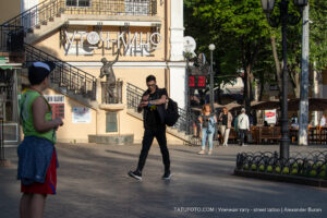 Фотография мужчины с татуировкой на руке, идущего по улице. Мужчина одет в черную футболку и джинсы. На заднем плане видна лестница и статуя.