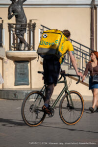 Фотография курьера Glovo на велосипеде с татуировкой на руке. Городской пейзаж, солнечный день, памятник на заднем плане.