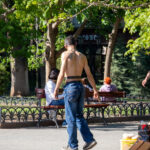 Фотография мужчины с татуировкой на спине, сидящего на скамейке в парке. Татуировка выполнена в черных тонах и имеет детализированный рисунок. На заднем плане деревья и другие люди.