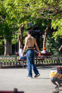 Фотография мужчины с татуировкой на спине, сидящего на скамейке в парке. Татуировка выполнена в черных тонах и имеет детализированный рисунок. На заднем плане деревья и другие люди.