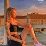 Фотография девушки с татуировкой в виде роз и черепа на руке, стоящей на фоне городского пейзажа.