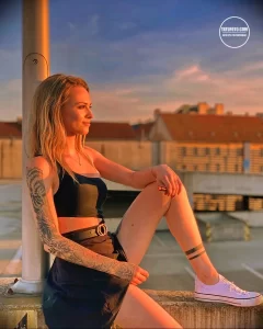 Фотография девушки с татуировкой в виде роз и черепа на руке, стоящей на фоне городского пейзажа.