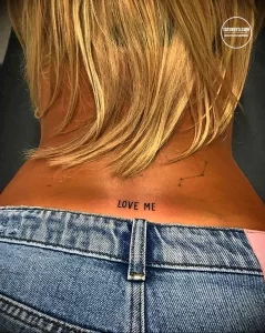Фото тату: Минималистичная надпись "LOVE ME" на женской спине, чуть ниже шеи. Небольшой эскиз татуировки, выполненный черными чернилами.