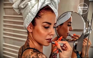 Фотография девушки с татуировкой на шее и руке. Девушка наносит макияж, отражаясь в зеркале.