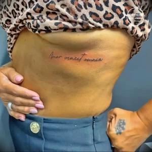 Фото татуировки с латинской надписью "Amor Vincit Omnia" (Любовь побеждает все) на боку девушки. Тату выполнено изящным шрифтом.