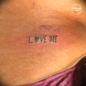 Фотография татуировки с надписью 'LOVE ME' и изображением сердца на груди. Рисунок выполнен черным цветом.