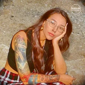 Фотография девушки в очках с татуировками на руках в стиле традишн, сидящей и задумчиво смотрящей в сторону.