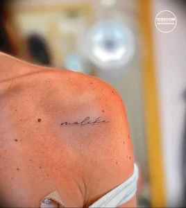 Фото тату с именем 'malika', выполненным простым шрифтом на плече девушки. Татуировка выглядит минималистично и элегантно.