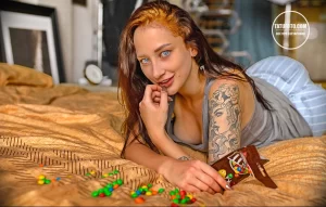 Фотография девушки с рыжими волосами, лежащей на кровати с татуировкой на плече и сладостями вокруг.