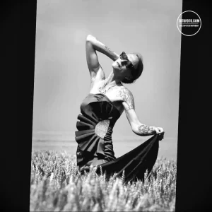 Черно-белое фото женщины в длинном платье и с татуировкой на плече, стоящей в поле пшеницы. Изображение подчеркивает красоту и грацию.