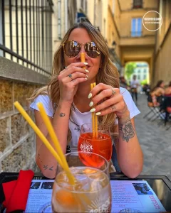 Фото девушки, сидящей за столиком с коктейлем Aperol Spritz и татуировками на руках. Видны рисунки цветов и абстрактных узоров. Подчеркивает стиль и любовь к отдыху.