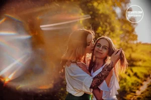 Фото тату на руке: рука девушки с татуировкой в виде ветви с цветами и листьями. Девушка обнимает другую девушку на фоне осеннего пейзажа.