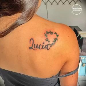 Фото тату на лопатке девушки: надпись 'Lucia' в красивом шрифте, обвитая цветами и листьями. Элегантный и женственный рисунок татуировки.