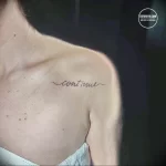 Фото минималистичной татуировки с надписью 'continue' на ключице девушки. Тонкие линии и лаконичный дизайн.