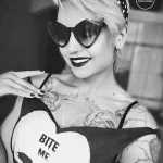 Фото тату черепа с надписью 'Bite Me' на плече девушки в ретро-стиле. Черно-белое изображение, акцент на татуировке и выразительном макияже.