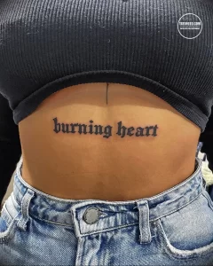 Фото тату на животе девушки: надпись "burning heart" готическим шрифтом, выполненная черным цветом. Девушка демонстрирует татуировку, стоя перед зеркалом.
