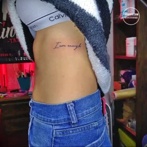 Фото минималистичной татуировки надписи "I am enough" на боку. Татуировка выполнена черным цветом и имеет простой шрифт.