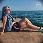 Фотография девушки, сидящей на берегу моря с татуировками на руках и ногах. Она выглядит расслабленной и счастливой, наслаждаясь видом на море.