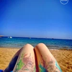Фото акварельной татуировки на ногах девушки на фоне моря и пляжа. Яркие цвета, абстрактные узоры. Девушка отдыхает на пляже.
