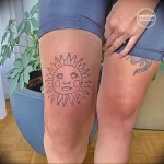 Фотография татуировки на колене, изображающей солнце с человеческим лицом. Оригинальный рисунок татуировки с четкими линиями и выразительным дизайном.