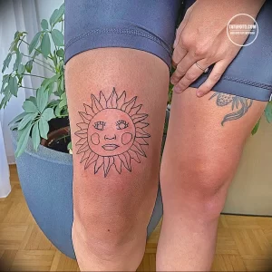 Фотография татуировки на колене, изображающей солнце с человеческим лицом. Оригинальный рисунок татуировки с четкими линиями и выразительным дизайном.