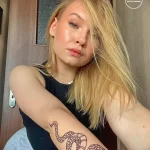 Фото татуировки змеи на руке девушки с длинными светлыми волосами. Рисунок выполнен тонкими линиями в красноватых тонах.