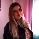 Фотография девушки с длинными светлыми волосами и татуировкой в виде геометрического узора на шее. Она смотрит в камеру с легкой улыбкой.
