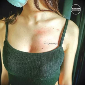Фото татуировки на груди с надписью 'Love yourself' и небольшим сердечком. Выполнено тонким шрифтом в черном цвете.