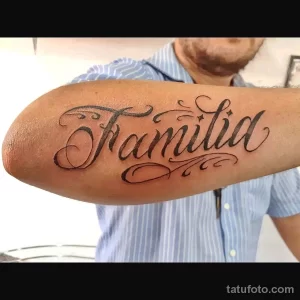 Фото классической татуировки надписи 'Familia' выполненной каллиграфическим шрифтом на предплечье мужчины. Черно-белый рисунок татуировки.