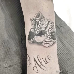 Фото тату на предплечье, изображающее детские кеды и имя "Alice". Детализированный рисунок татуировки в черно-белой гамме.