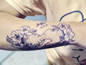 Фото тату на предплечье, изображающее детскую ручку, держащуюся за цветы. Детализированный рисунок татуировки в стиле графика.