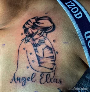 Фото тату на груди с изображением женщины, держащей ребенка. Надпись "Angel Elias" и маленькие сердечки дополняют композицию. Черно-белый рисунок.
