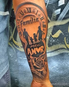 Фото тату на предплечье с надписью 'Familia' и силуэтами семьи, украшенными розой и римскими цифрами. Красивый рисунок татуировки для выражения семейных ценностей.